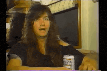a man with long hair is sitting at a table holding a can of bud light .