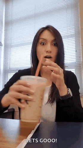 a woman is sitting at a table holding a cup of coffee with a straw .