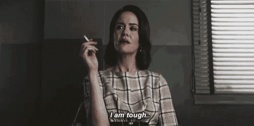a woman is smoking a cigarette in front of a window and says `` i am tough '' .