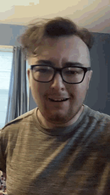 a man wearing glasses and a grey shirt smiles in front of a window