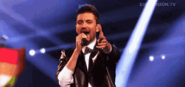 a man singing into a microphone on a stage with eurovision.tv in the background