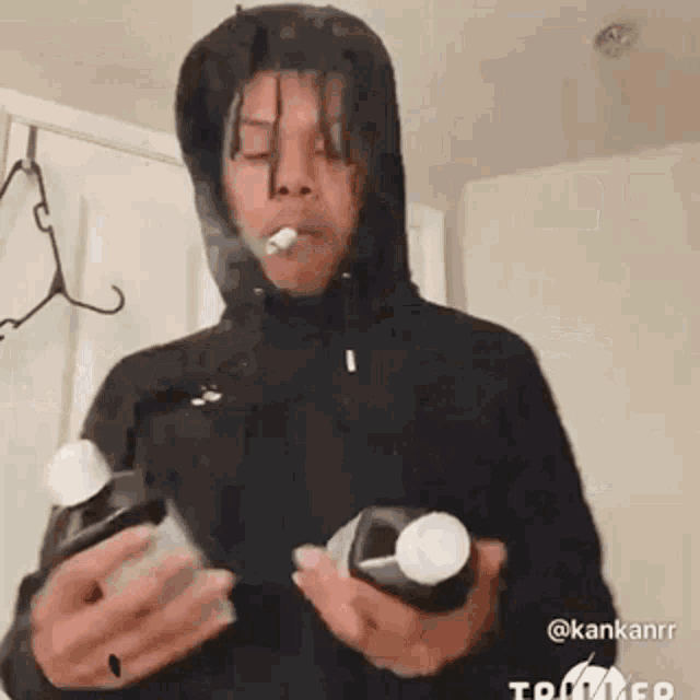 a man in a hoodie is smoking a cigarette while holding two bottles of pills .