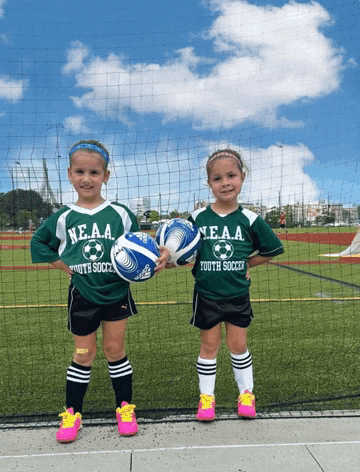 two young girls wearing neaa youth soccer jerseys stand on a field holding soccer balls