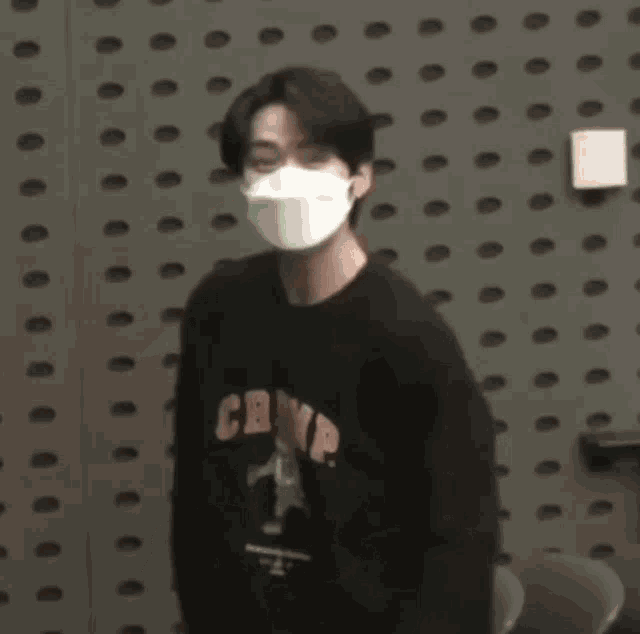 a man wearing a mask and a black sweater is standing in front of a wall with holes .