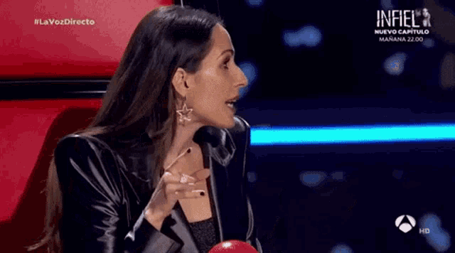 a woman in a black jacket is sitting in a chair on a stage and talking to someone .