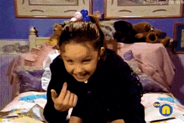 a little girl is sitting on a bed and giving the middle finger with a bbc logo in the corner