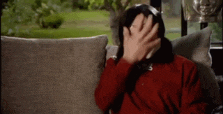 a man in a red shirt is sitting on a couch and covering his face with his hand .