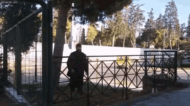 a man wearing a mask stands in front of a fence with the website greenof.com visible
