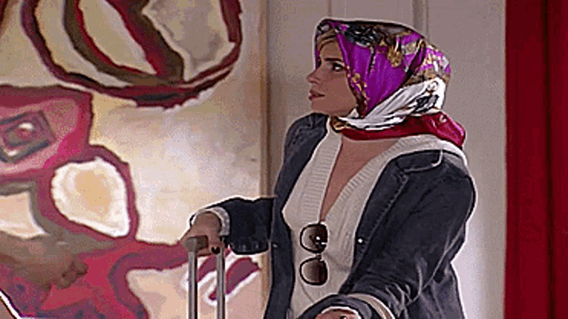 a woman wearing a scarf and sunglasses holds a suitcase in front of a painting