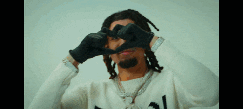 a man wearing a white sweater and black gloves makes a heart shape with his hands