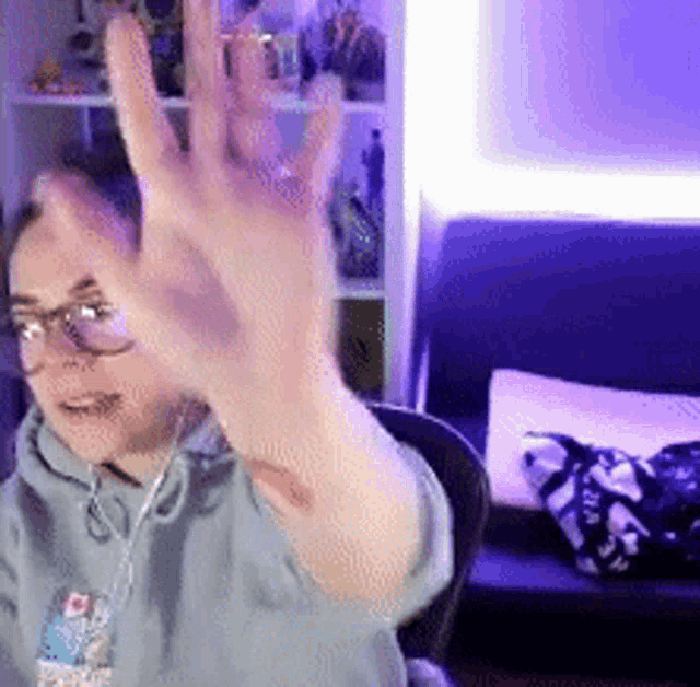 a person wearing glasses and a hoodie is waving their hand in front of a couch .