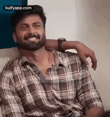 a man with a beard wearing a plaid shirt is sitting on a couch and smiling .