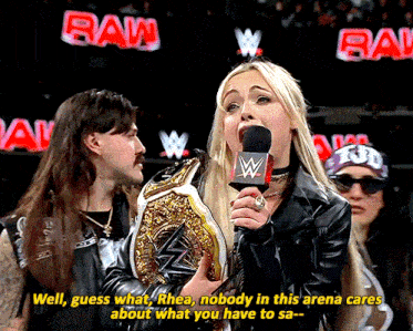 a woman holding a wrestling championship talks into a microphone