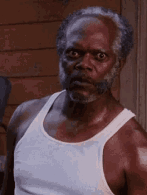 an older man with a beard is wearing a white tank top and looking at the camera .