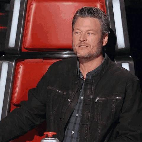 a man in a black jacket is sitting in a red chair with a red button .
