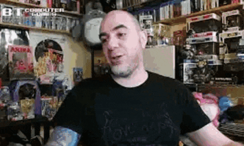a bald man with a tattoo on his arm is sitting in front of a shelf full of toys .