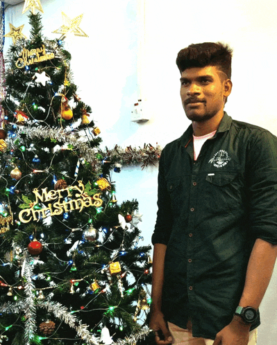 a man standing in front of a christmas tree with a merry christmas sign