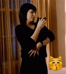 a woman in a black shirt is standing next to a yellow cat with a kissing face .