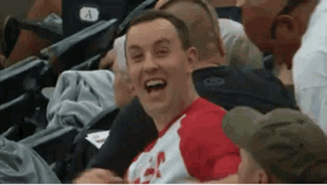 a man in a red shirt is laughing in a crowd of people at a baseball game .