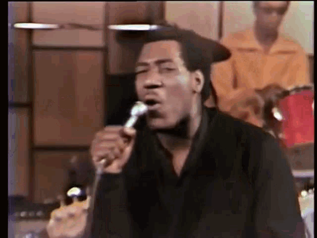 a man is singing into a microphone while a band plays drums in the background .