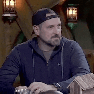 a man with a beard and a baseball cap is sitting at a table with a cup and a miniature house .