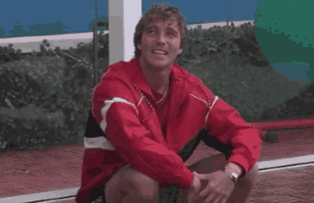 a man in a red jacket is making a heart shape with his hands while sitting on the ground .
