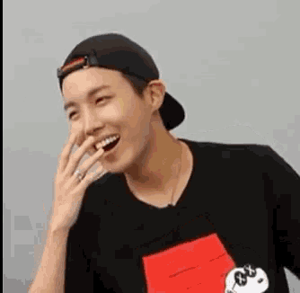 a man wearing a baseball cap and a black shirt is laughing while covering his mouth with his hand .