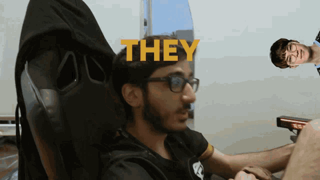 a man with glasses sits in a chair with the word they behind him