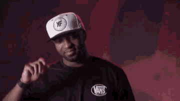 a man wearing a black vans shirt and a white hat is standing in front of a red wall .
