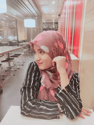 a woman in a hijab sits at a table with her hand on her chin