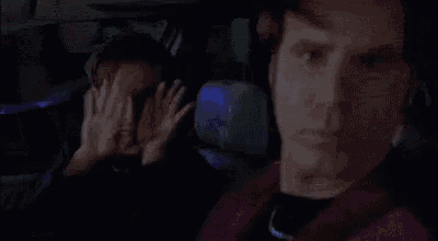 two men are sitting in a car one is covering his mouth with his hand .