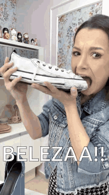 a woman in a denim jacket is holding a white shoe in her mouth with the words belezaa written on the bottom
