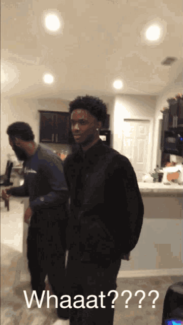 a man in a black jacket is standing in a kitchen with the words whaaat written below him