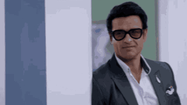 a man wearing glasses and a suit leans against a wall