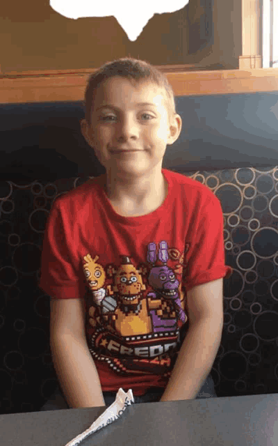 a young boy wearing a red five nights at freddy 's shirt sits at a table