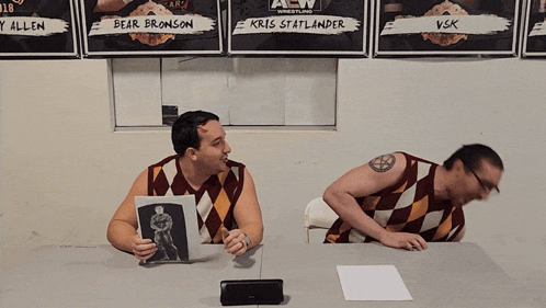 bear bronson and kris statlander sit at a table in front of a wall of wrestling posters