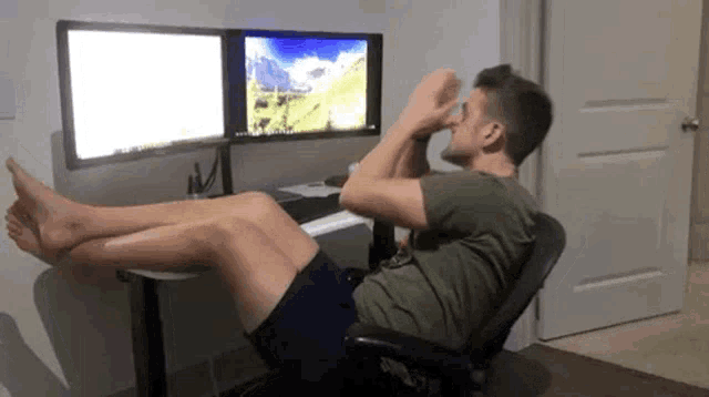 a man is doing crunches in front of two computer monitors while sitting in an office chair .