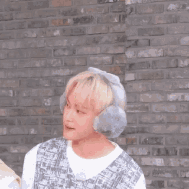 a young boy wearing ear muffs and a bow on his head is standing in front of a brick wall .