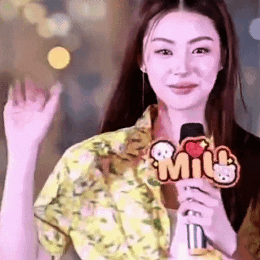 a woman is holding a microphone and waving at the camera while wearing a yellow shirt .