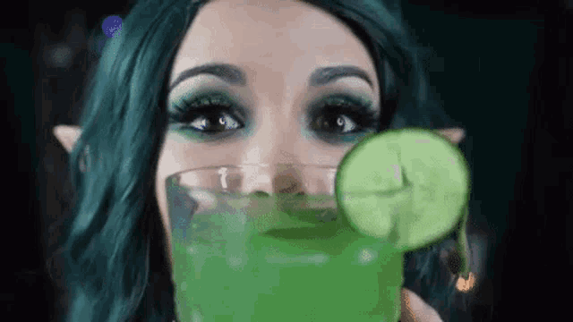 a woman in a green wig is drinking a green drink with a cucumber slice in it .