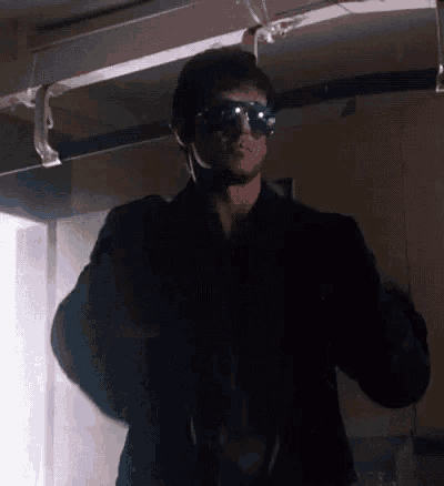 a man wearing sunglasses and a jacket is standing in a dark room .