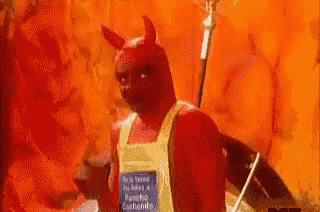 a man in a devil costume is holding a glass of wine in front of a wall of fire .
