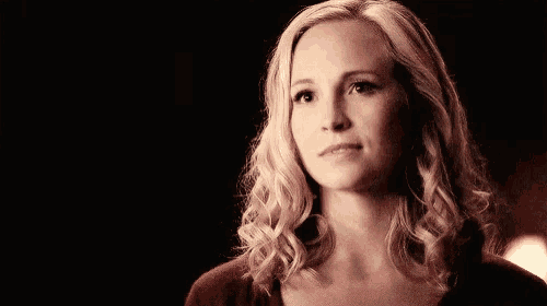 a woman with blonde curly hair and a red shirt is looking at the camera in a dark room .
