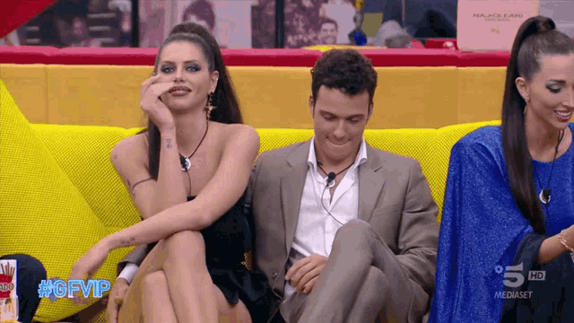 a man in a suit sits on a yellow couch next to a woman in a black dress and a woman in a blue dress
