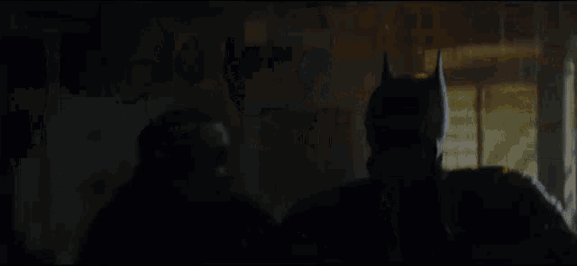 a man in a batman mask is standing in a dark room with another man .