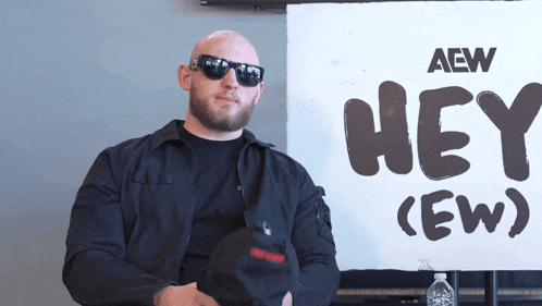 a man wearing sunglasses sits in front of a sign that says hey