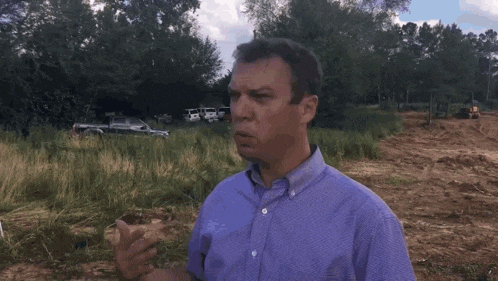a man in a purple shirt is standing in a field talking to a camera .