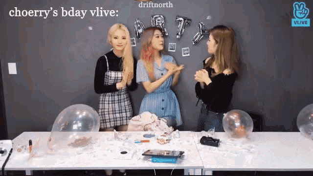 three girls are sitting at a table with balloons and a sign that says choerry 's bday vlive .