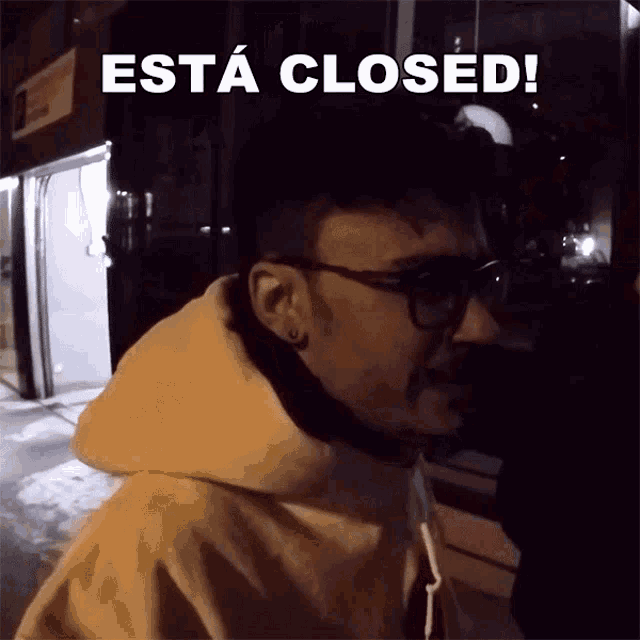 a man wearing glasses and a hoodie is standing in front of a sign that says " esta closed "