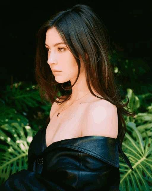 a woman wearing a black leather jacket is standing in front of a green plant